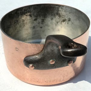 Peut inclure: Une casserole ronde en cuivre avec une poignée en fonte sombre fixée par trois rivets en cuivre. L'extérieur est en cuivre poli, tandis que l'intérieur présente un revêtement métallique avec une décoloration sombre et tachetée.