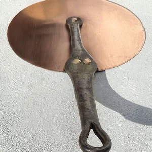 May include: A copper-colored, round lid with a riveted, dark metal handle. The handle has a loop at the end for hanging. The lid's surface has a slight sheen, and the handle shows signs of age.