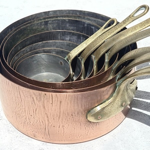 Pode incluir: Um conjunto de panelas de cobre com alças de latão. As panelas estão encaixadas umas nas outras, mostrando seus tamanhos variados. O exterior de cobre tem um tom marrom-avermelhado quente, enquanto os interiores mostram uma pátina mais escura.