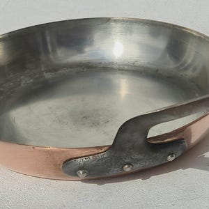 Könnte beinhalten: Eine runde Kupferpfanne mit einer polierten, silberfarbenen Innenbeschichtung. Die Außenseite hat ein warmes Kupferfinish und verfügt über einen dunklen, robusten Metallgriff, der mit drei sichtbaren Nieten an der Seite befestigt ist.
