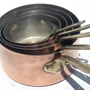 Könnte beinhalten: Ein Set aus fünf ineinander stapelbaren Kupfertöpfen mit silberfarbenen Metallgriffen. Jeder Topf hat eine glatte Kupferaußenseite und ein metallisches Inneres. Der größte Topf trägt die Prägung J.JACQUOTOT 77. RUE DAMREMONT PARIS MADE IN FRANCE.
