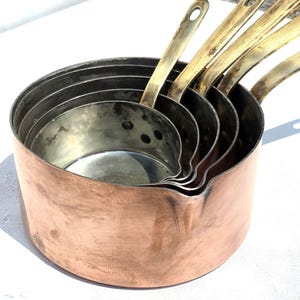 May include: A set of four nested copper saucepans with silver-toned interiors and long, slender brass handles. Each pan features a rounded body with a pouring spout on the rim, showcasing a metallic finish with visible signs of use and patina.