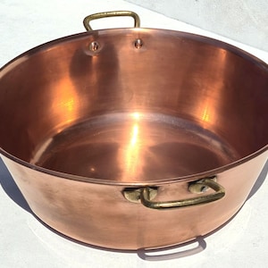 Puede incluir: Un cuenco grande y redondo de cobre pulido con una superficie interior y exterior lisa. El recipiente cuenta con dos asas de latón curvas y resistentes, fijadas de forma segura a los lados opuestos con remaches visibles para facilitar el agarre.