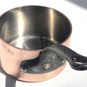 Puede incluir: Una cacerola de cobre con interior de acero inoxidable y un asa oscura. La cacerola tiene forma redondeada y un asa pequeña remachada. El exterior de cobre tiene un tono marrón rojizo cálido.