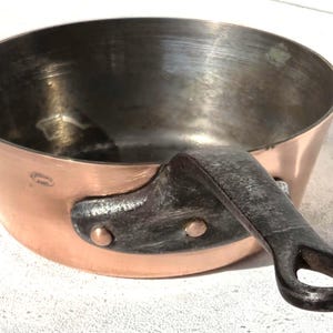 May include: A copper saucepan with a dark metal handle. The pan has a rounded shape with a shiny interior and a copper exterior. The handle is attached with rivets. A classic kitchenware item.