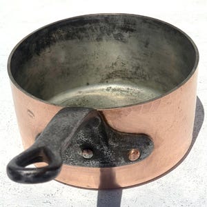 May include: A copper saucepan with a hammered finish and a dark metal handle. The interior is a lighter metal, showing signs of use. The handle is riveted to the side of the pan.