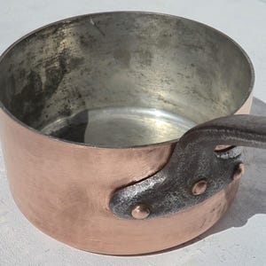 Pode incluir: Uma panela redonda de cobre com exterior polido e interior metálico com sinais de uso. A panela possui uma alça de ferro fundido escura e texturizada, fixada na lateral com três rebites de cobre visíveis, criando uma conexão firme.