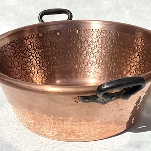 May include: A copper bowl with a hammered texture on the interior and a smooth exterior. The bowl has two black handles and a wide rim. The copper has a warm, reddish-brown color, and the bowl appears to be vintage.