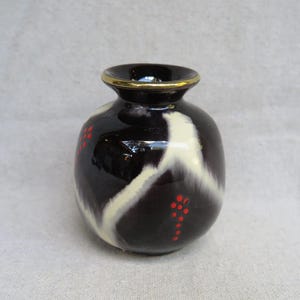 May include: A small, black ceramic vase with a gold rim. The vase features a white, abstract design and small red dot clusters. The vase is a decorative item.