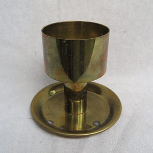 May include: A vintage brass candlestick holder with a circular base and a cylindrical cup. The candlestick holder has a warm, golden hue and shows signs of age. The design is simple and elegant, suitable for various decor styles.