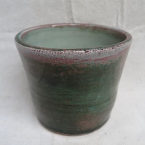 May include: A handmade ceramic tumbler with a green and brown glaze. The rim has a silver and red accent. The cup has a slightly flared shape and is suitable for beverages. The pottery has a rustic, organic aesthetic.
