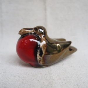 May include: A ceramic figurine of a robin bird. The bird has a glossy red breast, a brown head, and bronze-colored wings. The figurine is small and detailed, with a smooth, reflective surface.