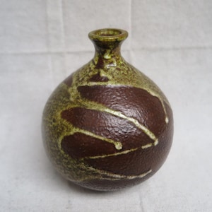 May include: A small, round ceramic vase with a narrow neck. The vase is primarily brown with a textured surface and is decorated with a pattern of olive green and white streaks. The vase is likely handmade.