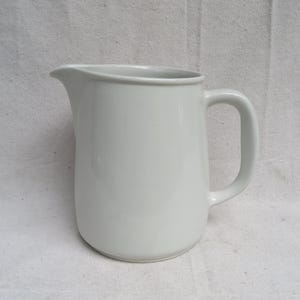 May include: A white ceramic pitcher with a curved handle and a pouring spout. The pitcher has a smooth, glossy finish and a simple, elegant design. It is likely used for serving beverages.