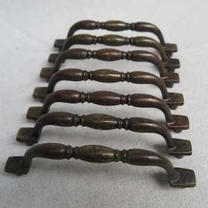 May include: A set of seven antique bronze drawer pulls. Each pull has a curved handle with a series of rounded sections and rectangular mounting plates. The bronze has a weathered, aged appearance, with variations in color.