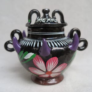 May include: A black ceramic vase with a lid, adorned with a floral design in pink and white, and accented with purple and green details. The vase has two handles and the word "MEXICO" is on the lid.