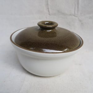 May include: A small, round ceramic bowl with a smooth white base and a contrasting dark olive-brown lid. The lid features a central circular knob handle and a glossy finish, while the bowl has a simple, clean silhouette and a matching brown rim.