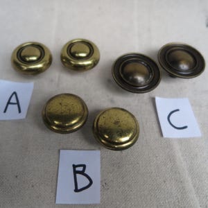 May include: A collection of six antique brass cabinet knobs. The knobs vary in design, with some featuring a raised center and others a flat, circular design. The brass has a warm, aged patina, suggesting vintage hardware.
