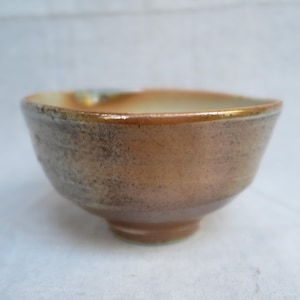 May include: A small, handcrafted ceramic bowl with a rounded shape and a footed base. The bowl features a mottled, earthy color palette of brown, tan, and gold, with a slightly uneven rim. The interior has a lighter, cream-colored glaze.