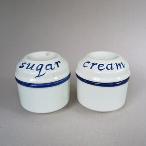 May include: Two white ceramic sugar and cream containers with blue trim and handwritten labels. The sugar container says "sugar" and the cream container says "cream".