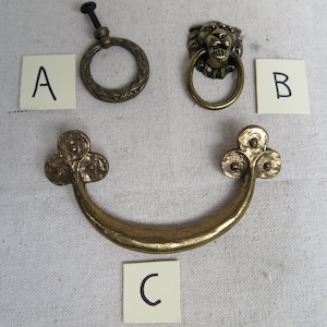 May include: Three antique brass hardware pieces: a ring pull with a vine design, a lion head ring pull, and a curved handle with decorative ends. Each piece is labeled with a letter on a white card.