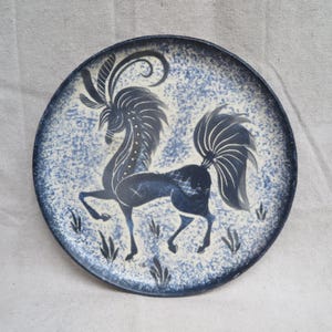May include: A round ceramic plate with a blue and white speckled background. A dark blue horse-like figure with a long tail and decorative head is in the center. The plate has a dark blue border and is approximately 10 inches in diameter.