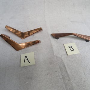 May include: Copper-colored cabinet hardware. Two sets of handles are shown. One set is shaped like a V, and the other is a curved bar. Each handle has a different design. The handles are labeled A and B.