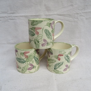 May include: Three white ceramic mugs with a green and purple floral pattern. The mugs are stacked on top of each other.