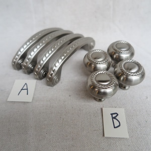 May include: A collection of silver-toned cabinet hardware, including four curved handles and four round knobs. The handles have a decorative rope-like edge, and the knobs feature a similar design. The items are arranged on a light-colored surface, with labels marked "A" and "B".