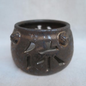May include: A small, bronze-colored ceramic container with cut-out Chinese characters. The container has a rounded shape with two small, raised decorative elements on the sides. The container is likely used for decorative purposes.