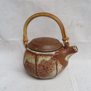 May include: A small, brown and white ceramic teapot with a bamboo handle. The teapot has a rounded body and a lid with a small hole in the top.