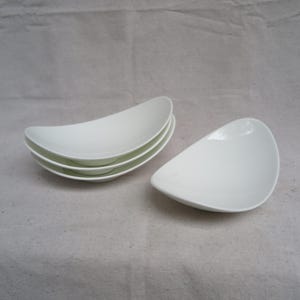 May include: Four white ceramic bowls stacked on top of each other. The bowls are oval shaped and have a slight curve to them. The bowls are on a white surface.