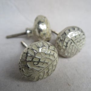 May include: A set of three silver-toned cabinet knobs with a textured, mosaic-like design. The knobs have a rounded shape with a stem for mounting. The mosaic pattern is a light cream color, creating a contrast with the silver metal.