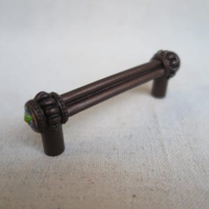 May include: A bronze-colored cabinet pull with decorative ends. The pull has a bar with two parallel rods and decorative ends with a circular design and a small, colorful jewel. The hardware is designed for furniture.