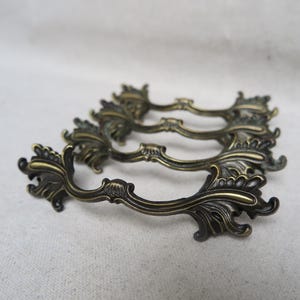 May include: Ornate antique brass drawer pulls. Each pull features a curved handle with decorative floral and leaf detailing. The pulls are arranged in a group, showcasing their intricate design and aged finish. Ideal for vintage furniture restoration.