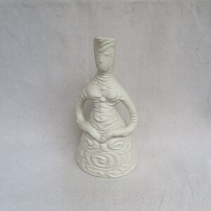 May include: White ceramic sculpture of a stylized woman with a textured surface. The sculpture is in a standing pose with arms crossed in front of her.