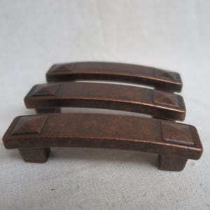 May include: Three bronze-colored cabinet pulls with a rectangular design and square accents on each end. The pulls have a curved shape and are made of metal. The hardware is suitable for drawers or cabinets.