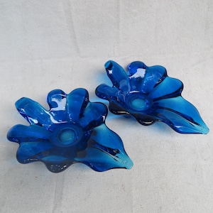 May include: Two cobalt blue glass flower-shaped bowls. Each bowl has a central circular indentation and petal-like edges. The glass has a glossy finish, with darker blue accents.