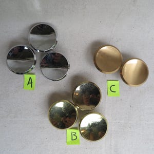 May include: An assortment of metallic, circular objects. Three are silver, three are gold, and two are brass-colored. Each has a slightly domed surface. The objects are labeled A, B, and C on small green sticky notes.