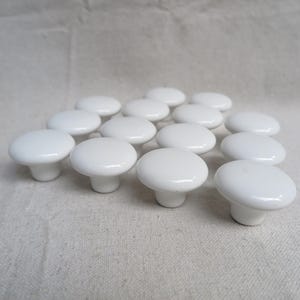 May include: A collection of fourteen white ceramic cabinet knobs arranged in a group. Each knob features a smooth, rounded top surface and a cylindrical base, providing a uniform appearance across the entire set of hardware components.