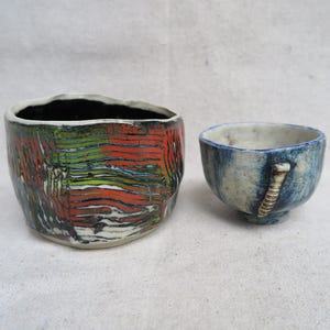 May include: Two handmade ceramic vessels. The larger one is a textured, cylindrical bowl with a black interior and a colorful exterior featuring red, green, and white hues. The smaller vessel is a blue cup with a handle.