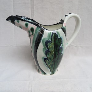 May include: A white ceramic pitcher with a unique spout and handle. The pitcher is decorated with green, black, and pink floral patterns.