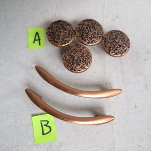 May include: A collection of copper-toned cabinet hardware. The set includes four round knobs with a textured, dark pattern and two curved handles. The hardware is displayed on a light-colored surface.