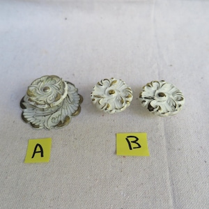 May include: A collection of white and gold floral cabinet knobs and pulls. The hardware features a detailed, ornate design with a distressed finish. The set includes a pair of knobs and a larger pull, perfect for adding a vintage touch to furniture.