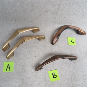 May include: Four vintage cabinet handles in gold and bronze tones. Two gold handles are at the top left, and two bronze handles are at the bottom right. Each handle has a curved, angular design, with a label next to each handle.