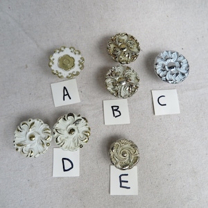 May include: A collection of decorative cabinet knobs in various designs and finishes. The knobs include floral and ornate patterns in white, gold, and silver tones. Each knob is labeled with a letter.