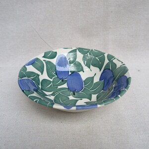 May include: A ceramic bowl with a white interior and a decorative exterior. The bowl features a pattern of green leaves and blue plums. The rim of the bowl is a light blue color. The bowl is oval-shaped.