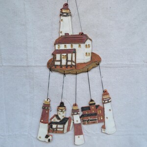 May include: A ceramic mobile featuring a lighthouse and several miniature buildings. The lighthouse is white with a brown top, and the buildings have brown roofs and tan walls. The mobile hangs from black strings.