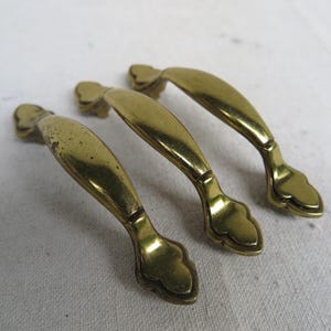 May include: Three vintage brass drawer pulls with a curved design and decorative ends. The pulls have a warm, golden hue and show signs of age, adding to their antique charm. These hardware pieces are ideal for restoring furniture.