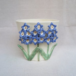 May include: A white ceramic planter with three blue and white flowers with gold centers. The flowers are in relief and have green leaves.
